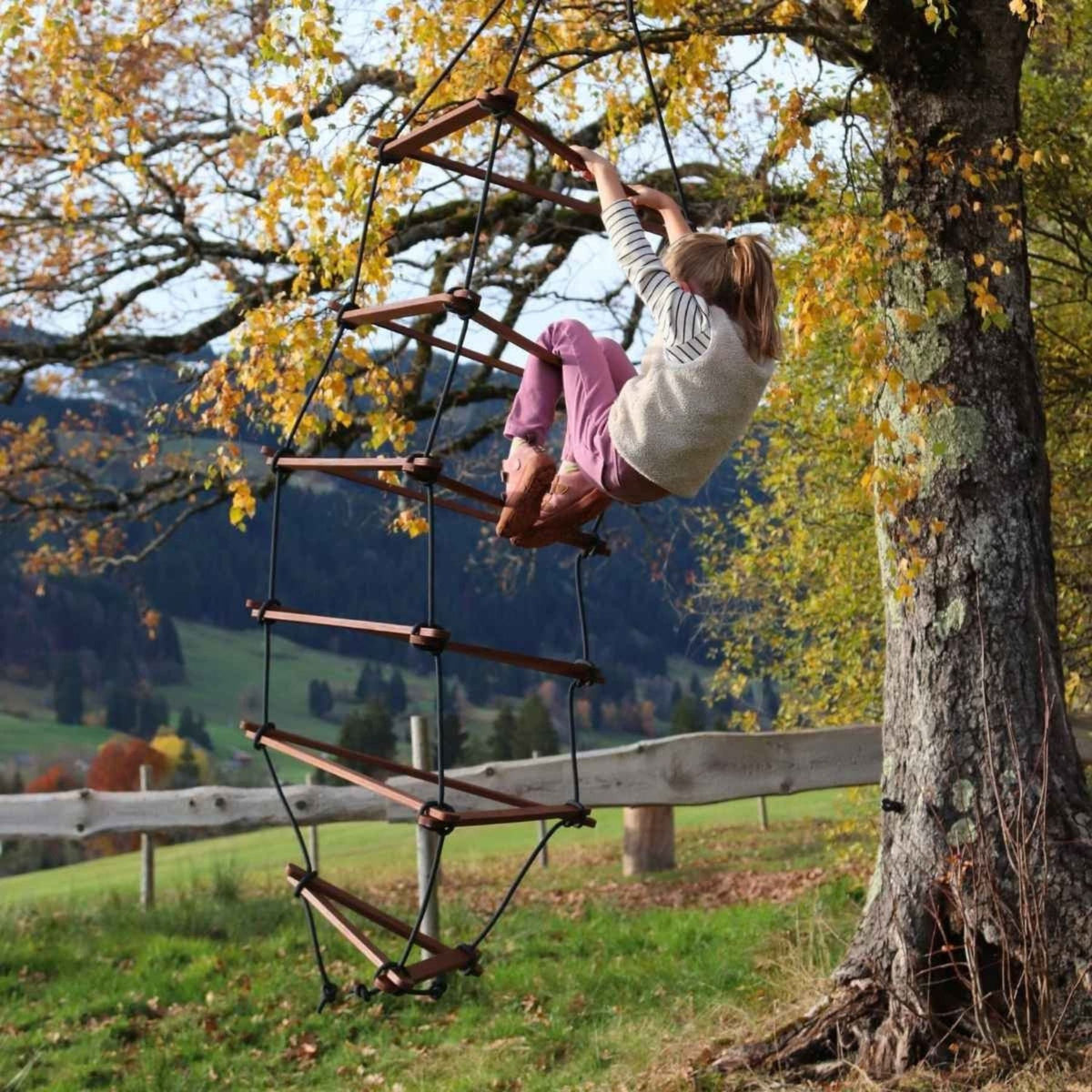 Mädchen klettert an der Strickleiter an einem Baum mit Herbstlaub, dahinter Hügel